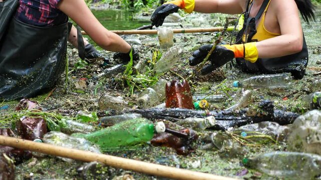 Female hands of eco activists in protective gloves cleaning messy river of debris. Unrecognizable volunteers in rubber suits collecting plastic bottles in dirty lake. Environment pollution protection