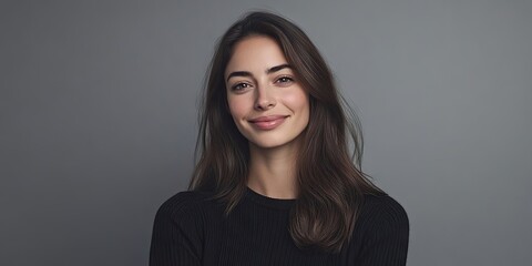 Naklejka premium Smiling brunette woman in a black sweater against a soft gray background expressing happiness and self-assurance with natural lighting.