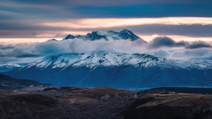 Mountain landscape under moody icy clouds with shades of blue and gray, snow-capped peaks emerging amidst low-hanging clouds at dusk.