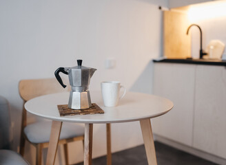 retro aluminum geyser coffee maker from Italy stands on a round beige table against the background of a kitchen with warm light.