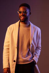 Fototapeta premium Stylish young man with sunglasses wearing a beige coat and a gray sweater, smiling against a colorful gradient background, embodying confidence and modern fashion
