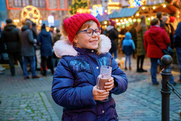 Little cute preschool girl drinking hot children punch or chocolate on German Christmas market. Happy child on traditional family market in Germany, Laughing kid in colorful winter clothes