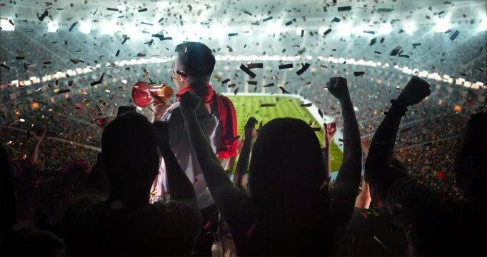 4K video of a lively stadium crowd at a nighttime soccer game, cheering with a large flag and confetti under bright lights