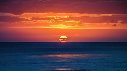 Obraz premium Vibrant sunset over ocean with deep orange and blue hues creating a serene atmosphere, low horizon with soft reflective waves in foreground.