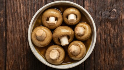 Pickled mushrooms in a ceramic bowl filled with brown liquid, arranged in a circle on a dark weathered wooden surface.