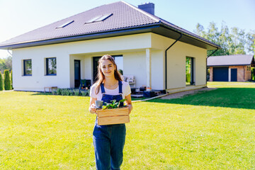 Young woman gardener wears jeans overalls standing in lawn holding box with houseplants and drinking coffee with her house on background.