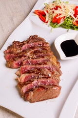 Costata di carne con cottura al sangue, scaloppata e servita in una braceria su un piatto bianco e accompagnata da salsa bbq e insalata con pomodori freschi e scaglie di formaggio