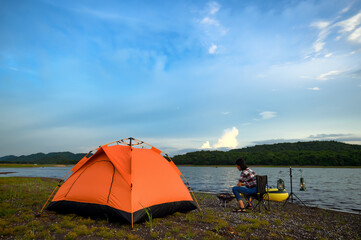 Orange tent set up by a peaceful lakeside, with a person sitting on a chair, enjoying the view under a bright blue sky.