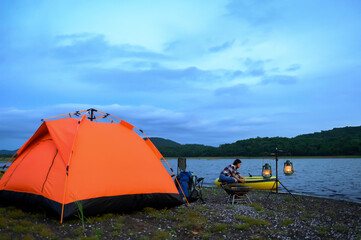 Orange tent set up by a tranquil lakeside, with a person preparing food near lanterns and a yellow kayak under a serene evening sky.