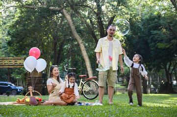 Cheerful family spends quality time in the park. Father blows giant bubbles while the kids play and...