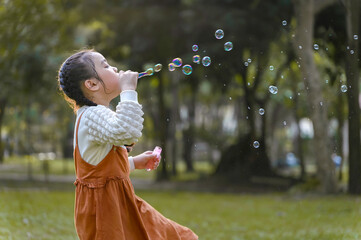 Young girl is blowing bubbles in a park. The scene is lighthearted and playful, with the girl...