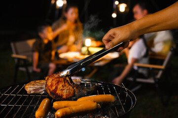 Family enjoys a nighttime barbecue outdoors. The focus is on a hand grilling meat and sausages, while the family sits in the background, sharing a cozy, festive meal under warm lights