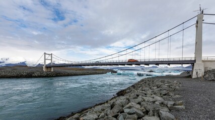 Suspension bridge over Jokulsarlon Glacier Lagoon in Iceland with icy waters and a cloudy sky.