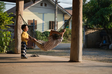 A cheerful boy interacts with his father resting on a hammock under a wooden porch in a rural Thai setting during sunset.