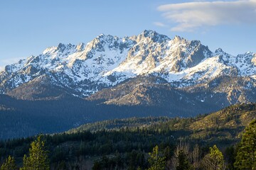 Fototapeta premium Majestic snow-capped mountains rise above lush green forests under a clear blue sky at sunset