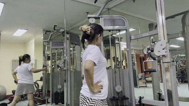 A woman is using exercise equipment at the gym. She is wearing a white t-shirt and a black and white pattern skirt. There are mirrors and other equipment in view.