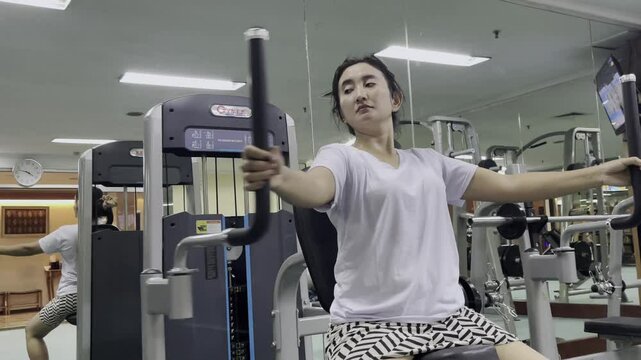 A person with a white shirt and black and white shorts uses a gym machine, the person is in a gym full of mirrors and exercise equipment.