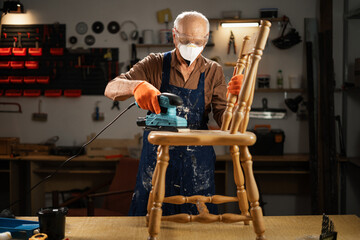 Obraz premium Senior man carpenter grinding wood chair with sandpaper in diy workshop. Electric sander machine working in carpentry.