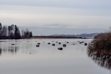  A flooded field with hay bales partially submerged, surrounded by trees and distant hills under an overcast sky, reflecting a serene atmosphere.