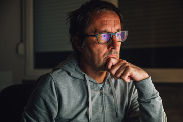 Portrait of adult caucasian freelancer working remotely from home late at night, his face with eyeglasses lit by blue screen light in low key environment of home office