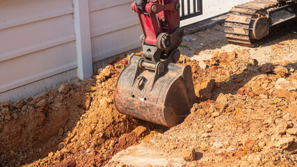 A construction excavator is digging a trench in the red earth for laying a drainage pipe in front of a house or building.