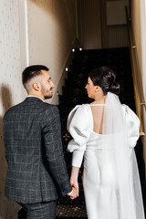 couple walking up the stairs. The man is wearing a gray checkered suit and a white shirt. Woman in a white dress with an open back and a long veil.
