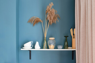 Wooden shelf in the kitchen with a vase of dry grass, a glass, tea cups and saucers, with a jar of cookies. Wooden shelf on a blue wall in a kitchen interior. Selective focus