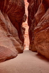 View of The Treasury (Al-Khazneh) through narrow Siq canyon in Petra, Jordan. Natural rock formations frame ancient architectural monument.