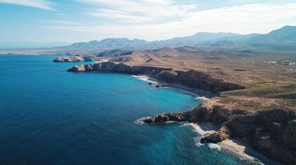 Fototapeta premium Aerial view of a rugged coastline with turquoise water meeting brown, arid hills and mountains under a bright blue sky