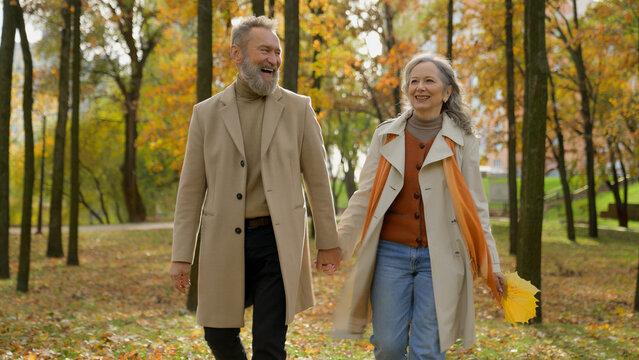 Happy old senior married couple adults hold hands together walking in city park joyful mature gray haired Caucasian woman wife and man husband have fun talking laughing walk stroll in autumn forest
