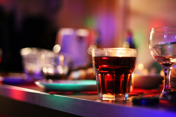 Full party table with dark red drink in a thick glass on the foreground. Colorful, bokeh background.