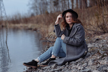 A girl in a polo and hat sits on the rocky shore of a lake at sunset, park, landscape, walk in the forest.