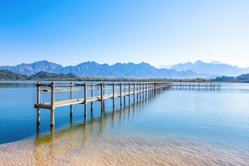 Obraz premium Serene wooden pier extending into calm waters, surrounded by majestic mountains under a clear sky