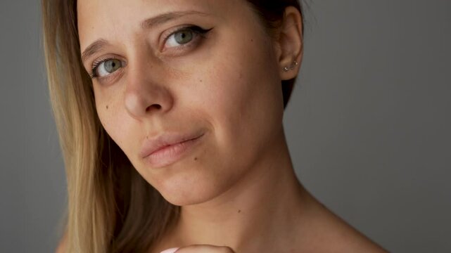 Young caucasian blonde woman showing the result of plastic surgery buccal fat removal stroking her cheek with her hand on a dark grey background. A female face with clear highlighted cheekbones