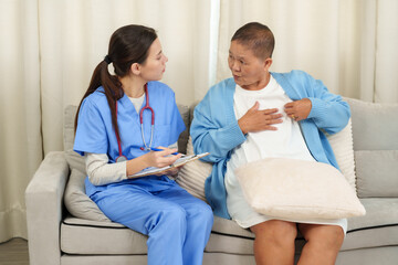 Young female nurse having a detailed discussion with elderly Asian woman during recovery session. Patient pointing to chest area while explaining symptoms in home healthcare setting.