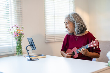 mature asian woman sitting on sofa looking tablet for practice music with ukulele in living room at...