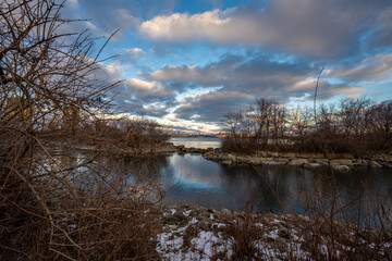 Obraz premium Winter's tranquil embrace on a waterway, where bare branches frame a distant city skyline under a dramatic sky. The scene is serene, a peaceful moment captured in nature's beauty.