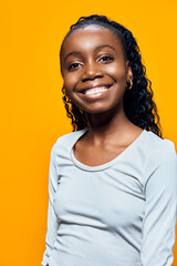 A cheerful young African girl with curly hair smiles brightly against a vibrant yellow background,...