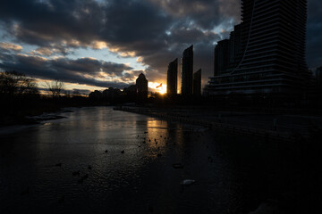 Fototapeta premium Silhouetted cityscape at sunset, reflecting in a calm river. Birds rest on the water's surface as the sun dips below the horizon, casting a warm glow on the clouds.