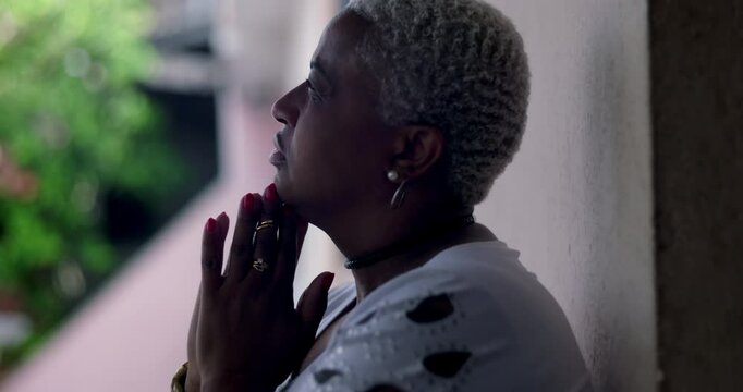 Woman in hopeful reflection, standing against wall, hands clasped in prayer, African descent, outdoor setting, facial expression conveying optimism