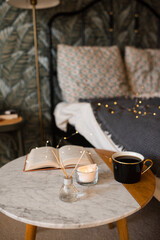 Marble coffee table with aromatic diffuser, open paper book, scented candle and cup of tea over bed in room. Cozy home atmosphere.