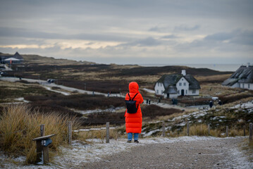Person in orange coat stands amidst winter dunes with frosty grasses, muted cloudy sky and distant thatched-roof houses on island Sylt, Schleswig-Holstein, Germany.
