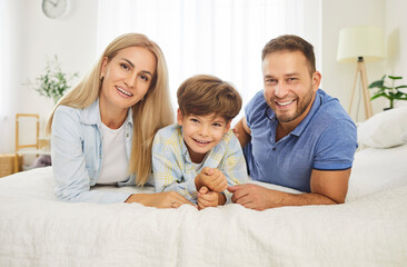 Fototapeta premium Portrait of young happy family of three lying on bed in bedroom with their child boy. Smiling parents - mother, father and son looking cheerful at camera resting at home on weekend.