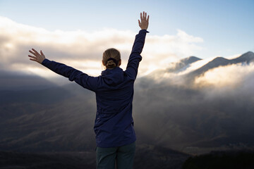 Backside view portrait of an unrecognizable woman in hiking wear with raised hands on top of the mountain in the sunset. Hiker girl with hands up. Feeling of freedom. Win, achieving success outdoors.