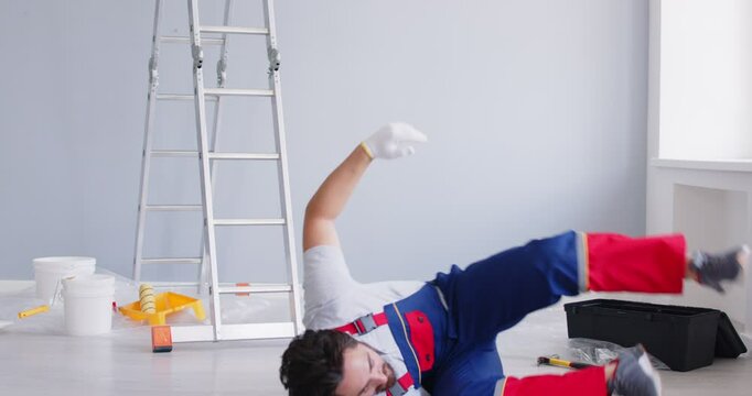 Worker handyman in overalls and safety hardhat doing home interior renovation starts climbing ladder, stumbles and falls down on floor with paint buckets, tools and equipment. Accident at work concept