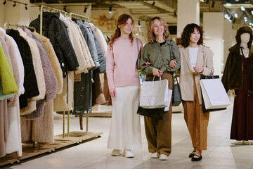 Wide shot of young adult customers with paper bags in hands looking at stands with new collection of female fur coats