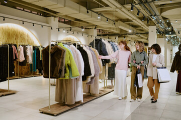 Company of three attractive girls with paper bags in hands looking at new collection of fur coats,...
