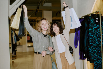 Portrait of biracial sisters hugging each other and raising hands with paper bags while shopping in...