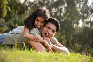 Fototapeta premium Indian happy father and daughter lying down and embracing, spending leisure time by looking at camera outdoors at park. Concept of indian father and daughter love