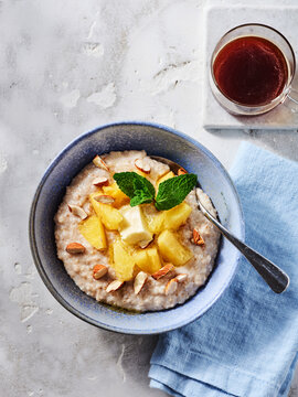 A bowl of creamy oatmeal is served with fresh pineapple chunks and sliced almonds, garnished with mint leaves. A small cup of tea sits beside it, creating a warm atmosphere